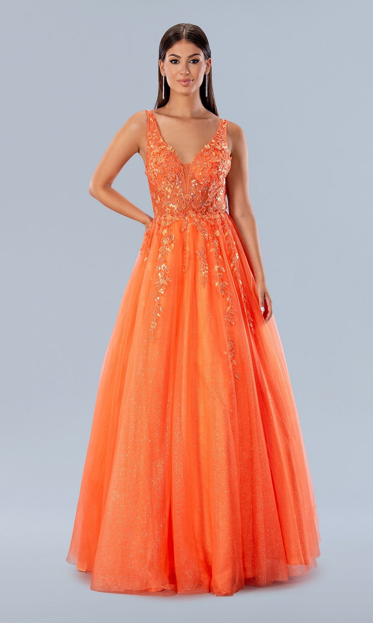 A woman poses in the Stella Couture 24203, a long glitter prom gown featuring a sleeveless, bright orange floral-embroidered bodice and a full sparkling tulle skirt, set against a plain background.